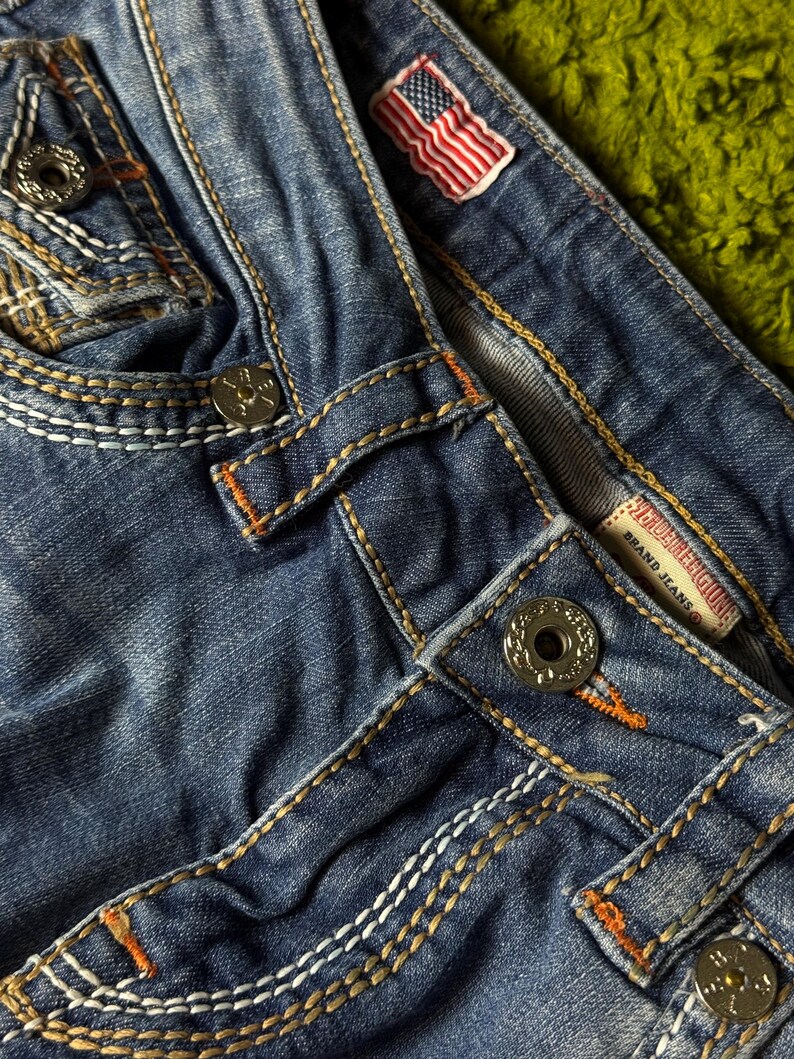 May include: Close-up of blue denim jeans with white and orange stitching. Features include metal buttons, a small American flag patch, and a brand label. The jeans have a worn, distressed look.