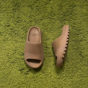 K&ouml;nnte beinhalten: Zwei beigefarbene Schlappen liegen auf einer gr&uuml;nen, strukturierten Oberfl&auml;che. Eine Sandale steht aufrecht und zeigt das offene Zehendesign, w&auml;hrend die andere auf der Seite liegt und die geriffelte Sohle zeigt. Die Sandalen scheinen aus weichem Material zu sein.