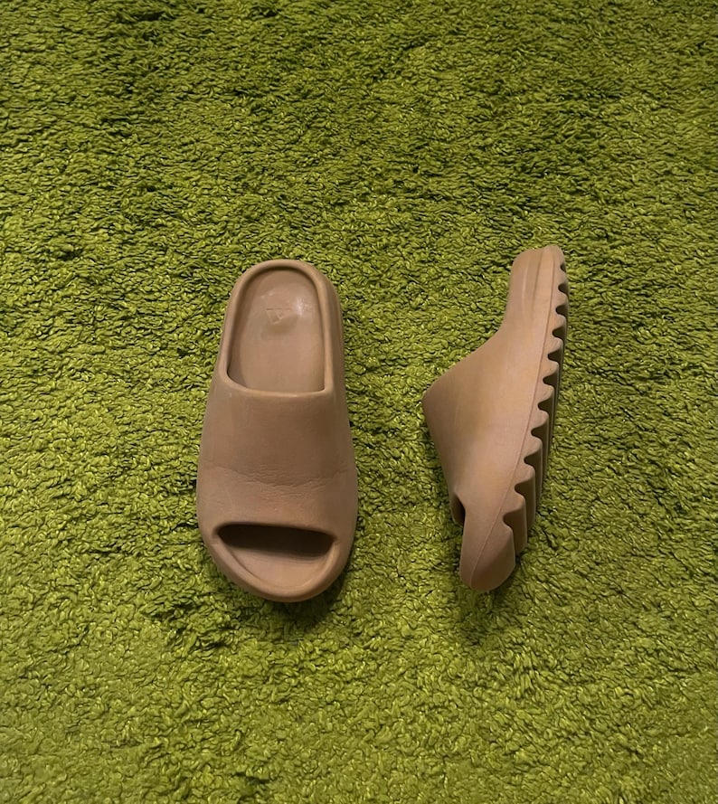 K&ouml;nnte beinhalten: Zwei beigefarbene Schlappen liegen auf einer gr&uuml;nen, strukturierten Oberfl&auml;che. Eine Sandale steht aufrecht und zeigt das offene Zehendesign, w&auml;hrend die andere auf der Seite liegt und die geriffelte Sohle zeigt. Die Sandalen scheinen aus weichem Material zu sein.