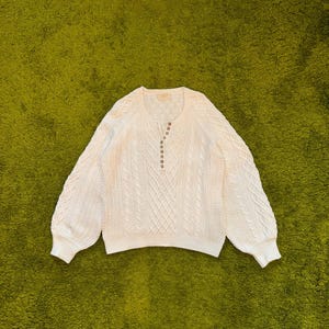 May include: A cream-colored long-sleeve sweater featuring a cable knit pattern, a round neckline, and a partial button-down placket at the center front. The garment has ribbed cuffs and a ribbed hem, with long sleeves and a textured knit construction.