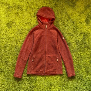 May include: A brick-red fleece jacket with a hood and a full-length zip. The jacket features two zipped pockets and a small logo on the upper left sleeve. The hood's interior is a lighter shade of red.