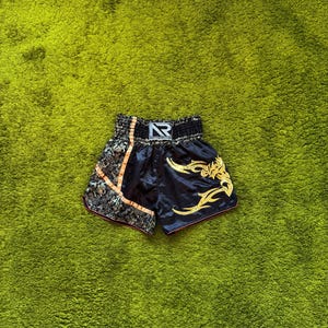 Puede incluir: Pantalones cortos de Muay Thai negros con diseño tribal dorado y un panel lateral decorativo estampado. La cintura presenta el texto "NOTHER BOXER" y un logotipo "NR" estilizado. Los pantalones cortos están sobre una superficie verde.