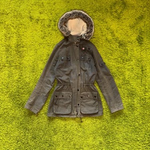 May include: An olive green jacket with a fur-trimmed hood. The jacket features multiple pockets, a drawstring waist, and a button-up front. The hood is lined with faux fur. The jacket is displayed on a green surface.