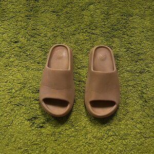 K&ouml;nnte beinhalten: Ein Paar beigefarbene Slides mit minimalistischem Design, einem einzelnen Riemen und offenem Zehenbereich. Die Slides bestehen aus einem glatten Material und befinden sich auf einem gr&uuml;nen, strukturierten Hintergrund.