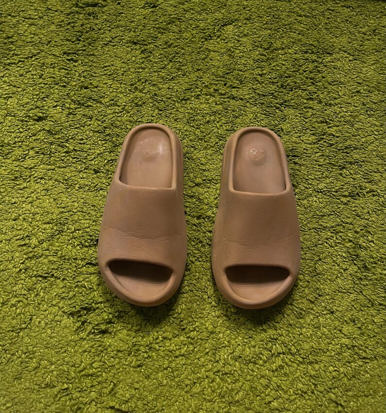 K&ouml;nnte beinhalten: Ein Paar beigefarbene Slides mit minimalistischem Design, einem einzelnen Riemen und offenem Zehenbereich. Die Slides bestehen aus einem glatten Material und befinden sich auf einem gr&uuml;nen, strukturierten Hintergrund.
