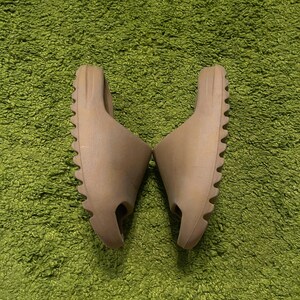 K&ouml;nnte beinhalten: Ein Paar hellbraune Slides mit strukturierter Sohle, platziert auf einem gr&uuml;nen, zotteligen Teppich. Die Slides haben ein minimalistisches Design mit einem einzelnen Riemen und einem konturierten Fu&szlig;bett, geeignet f&uuml;r den t&auml;glichen Gebrauch.