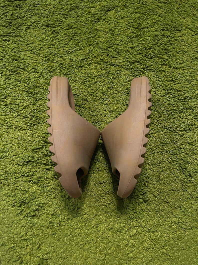 K&ouml;nnte beinhalten: Ein Paar hellbraune Slides mit strukturierter Sohle, platziert auf einem gr&uuml;nen, zotteligen Teppich. Die Slides haben ein minimalistisches Design mit einem einzelnen Riemen und einem konturierten Fu&szlig;bett, geeignet f&uuml;r den t&auml;glichen Gebrauch.