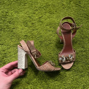 May include: A pair of tan woven fabric sandals with an ankle strap and a gold-tone buckle. The block heel features a white snakeskin-like texture. The front strap is embellished with a cluster of clear, faceted crystals in various shapes.