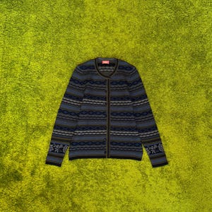 Pode incluir: Um cardigan azul e cinzento estampado com decote redondo e mangas compridas. O cardigan apresenta um design geométrico repetitivo e detalhes florais nos punhos. O fundo é verde brilhante.