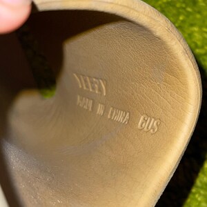 K&ouml;nnte beinhalten: Nahaufnahme einer beigefarbenen Slide-Sandale. Das Innere der Sandale ist sichtbar, mit dem Wort "YEEZY", "MADE IN CHINA" und "GUS" in Schwarz gedruckt.