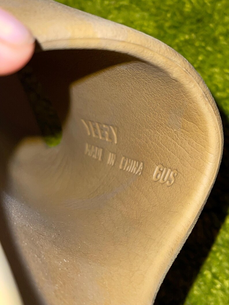 K&ouml;nnte beinhalten: Nahaufnahme einer beigefarbenen Slide-Sandale. Das Innere der Sandale ist sichtbar, mit dem Wort "YEEZY", "MADE IN CHINA" und "GUS" in Schwarz gedruckt.