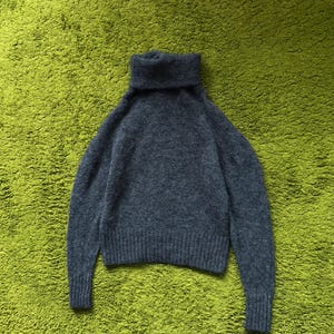 May include: A dark gray turtleneck sweater with long sleeves. The sweater is made of a soft, textured material and has a ribbed hem and cuffs. The turtleneck is folded over. The sweater is displayed on a green surface.