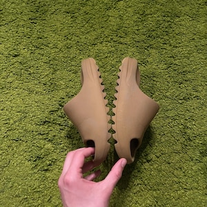 K&ouml;nnte beinhalten: Ein Paar hellbraune Schlupfsandalen. Die Sandalen haben ein geformtes Design mit strukturierter Sohle und einer einzelnen &Ouml;ffnung f&uuml;r die Zehen. Sie werden auf einer gr&uuml;nen, strukturierten Oberfl&auml;che pr&auml;sentiert.