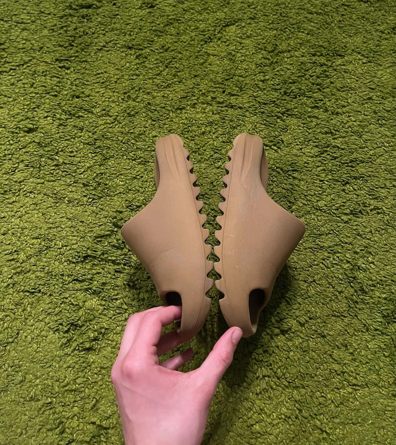 K&ouml;nnte beinhalten: Ein Paar hellbraune Schlupfsandalen. Die Sandalen haben ein geformtes Design mit strukturierter Sohle und einer einzelnen &Ouml;ffnung f&uuml;r die Zehen. Sie werden auf einer gr&uuml;nen, strukturierten Oberfl&auml;che pr&auml;sentiert.