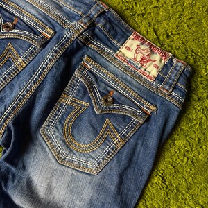May include: Close-up of blue denim jeans with white and gold stitching. The back pockets feature a distinctive horseshoe design. A red and white label is visible on the back, and the jeans are on a green surface.