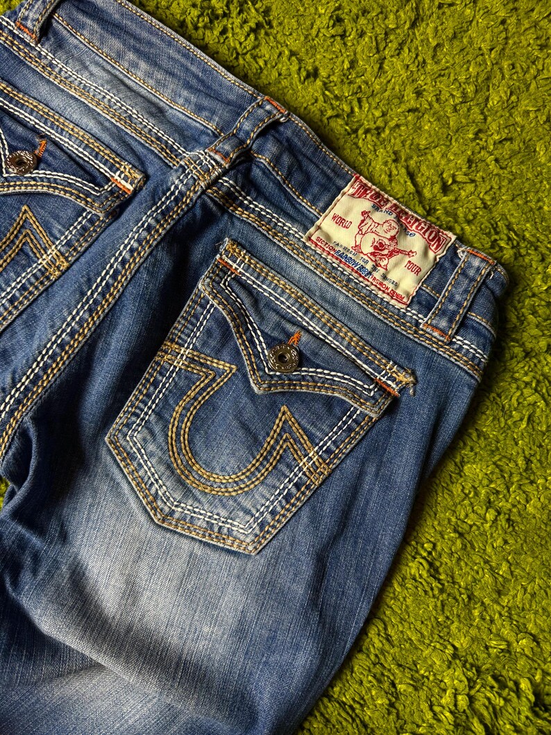 May include: Close-up of blue denim jeans with white and gold stitching. The back pockets feature a distinctive horseshoe design. A red and white label is visible on the back, and the jeans are on a green surface.