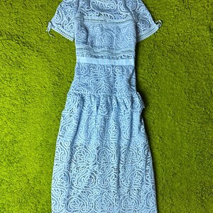 May include: A light blue lace dress with short sleeves and a midi length. The dress features intricate floral patterns throughout the fabric and a fitted waist. The dress is displayed on a green surface.