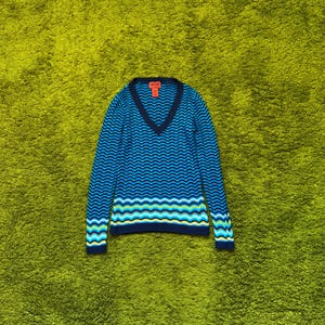 以下が含まれることがあります： 青いVネックセーター。青色の波模様が特徴です。長袖で、袖口と裾にはコントラストのあるトリムが付いています。ネックラインの近くに小さな赤いタグが見えます。