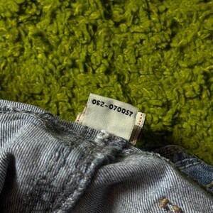 May include: Close-up of a section of blue denim fabric with a tag attached. The tag is white with the text "062-070057" printed in black. The background is a textured, bright green surface.