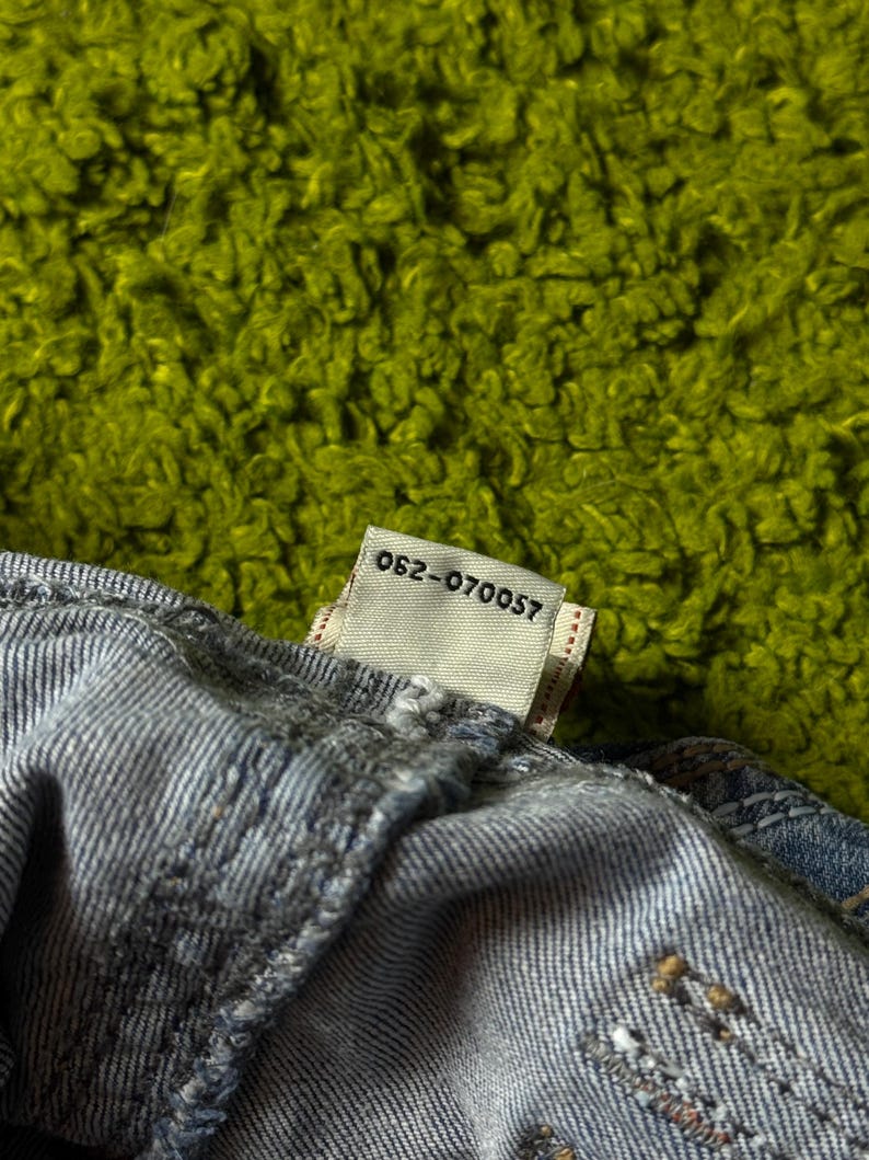 May include: Close-up of a section of blue denim fabric with a tag attached. The tag is white with the text "062-070057" printed in black. The background is a textured, bright green surface.