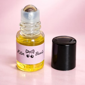 May include: A small, clear glass roll-on bottle filled with yellow liquid, next to a black cap. The bottle has a pink label with the text "Kitten Beauty" and a cherry illustration. The background is a soft pink.