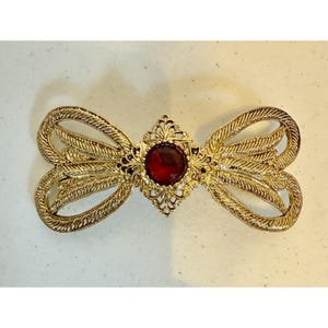 May include: A gold-toned bow-shaped brooch with a central, round, red gemstone. The brooch has an ornate, filigree design with textured detailing. The bow's loops are stylized and symmetrical, creating an elegant, vintage aesthetic.