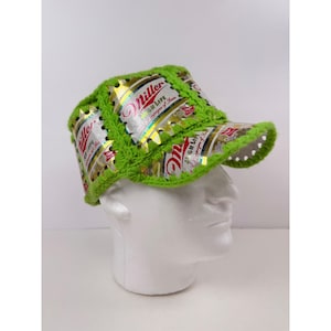 Puede incluir: Una gorra de béisbol construida con latas de cerveza Miller High Life aplanadas y unidas con tejido de ganchillo en hilo verde lima brillante. La gorra presenta una corona estructurada y una visera curva con el logo de Miller visible en el metal.