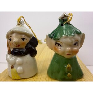 May include: Two ceramic bell ornaments with gold hanging strings. The left ornament is a white snowman figure with a black scarf and yellow broom. The right ornament is a green elf figure wearing a pointed hat with a star detail and gold buttons.