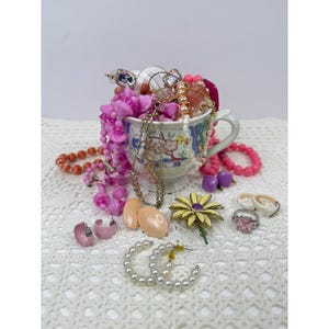 May include: A ceramic teacup filled with assorted jewelry including pink beaded necklaces, gold-tone chains, and pearl strands. Surrounding the cup are a yellow flower brooch, a pink stone ring, pearl hoop earrings, and various pink earrings.