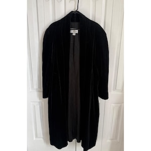 May include: A long, black velvet duster coat featuring a shawl collar, long sleeves, and an open front design. The interior is lined with a dark fabric, and a white brand label is visible at the inner neckline, displaying the text Collections.