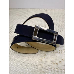 May include: A navy blue belt featuring a rectangular silver-tone metal buckle. The buckle is engraved with the text ESCADA. The belt has a smooth, light beige interior lining and a dark navy blue exterior strap with visible stitching.