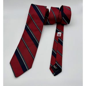 Puede incluir: Una corbata roja con rayas diagonales en azul marino, blanco y gris. La corbata está hecha de una tela texturizada y presenta una etiqueta con texto y una bandera británica. La corbata mide aproximadamente 147 cm de largo.