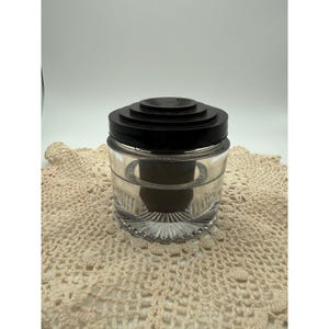May include: A clear glass jar with a thick, tiered black lid. The jar features a cylindrical body with a textured, ribbed base and a smooth, transparent upper section. A dark, solid cylindrical insert is visible inside the glass container.