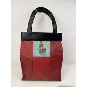 May include: A rectangular handbag featuring a patchwork design of deep red leather panels. A central light blue vertical panel displays a graphic illustration of a clown wearing a striped hat and a ruffled collar. The bag has a black leather top trim and handles.