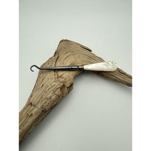 May include: A button hook featuring a dark metal shaft with a curved tip and a smooth, iridescent white mother-of-pearl handle. The handle is connected to the metal shaft by a small, ribbed brass-colored metal ferrule.
