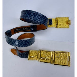 Puede incluir: Un cinturón de textura de piel de serpiente azul con una hebilla de metal dorado y tres placas rectangulares doradas con imágenes en relieve de caballos y jinetes. La etiqueta interior dice Alexis Kirk New York Paris London.