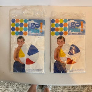 Op de afbeelding: Twee verpakkingen met strandballen. Elke verpakking toont een persoon die een strandbal vasthoudt met rode, gele, blauwe en witte panelen. De verpakking heeft de tekst "Gel for Sun" en een kleurrijk stippenpatroon.