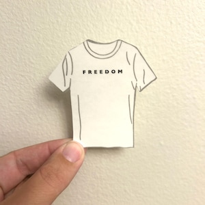 May include: A white t-shirt illustration with the word "FREEDOM" printed in black. The t-shirt is held by a hand against a neutral-colored wall. The design is simple and graphic.