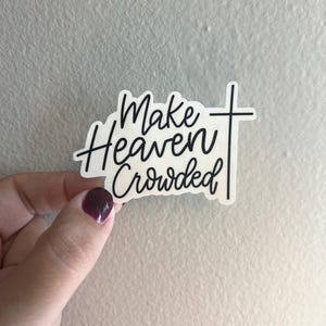 Op de afbeelding: Een witte sticker met zwarte tekst die "Make Heaven Crowded" leest in een handgeschreven lettertype. Een zwart kruis staat aan de rechterkant van de tekst. De sticker wordt vastgehouden door een hand met donkerrode nagellak.