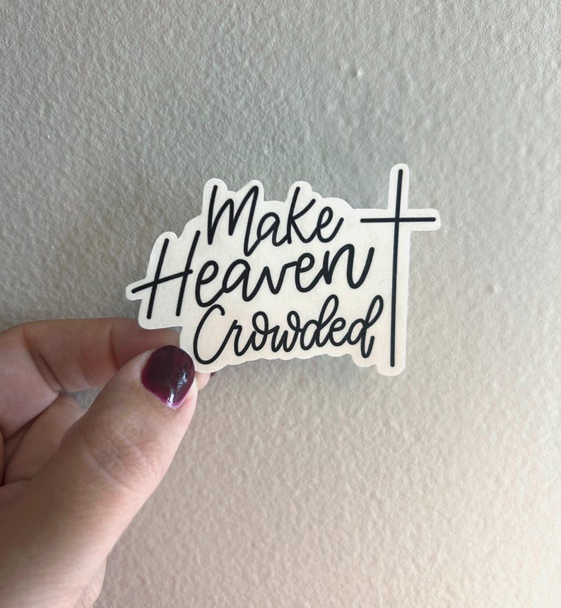 Op de afbeelding: Een witte sticker met zwarte tekst die "Make Heaven Crowded" leest in een handgeschreven lettertype. Een zwart kruis staat aan de rechterkant van de tekst. De sticker wordt vastgehouden door een hand met donkerrode nagellak.