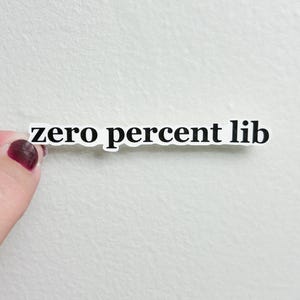 May include: A white sticker with the text "zero percent lib" in a black, sans-serif font. The sticker is held by a person with a light skin tone and dark red nail polish.