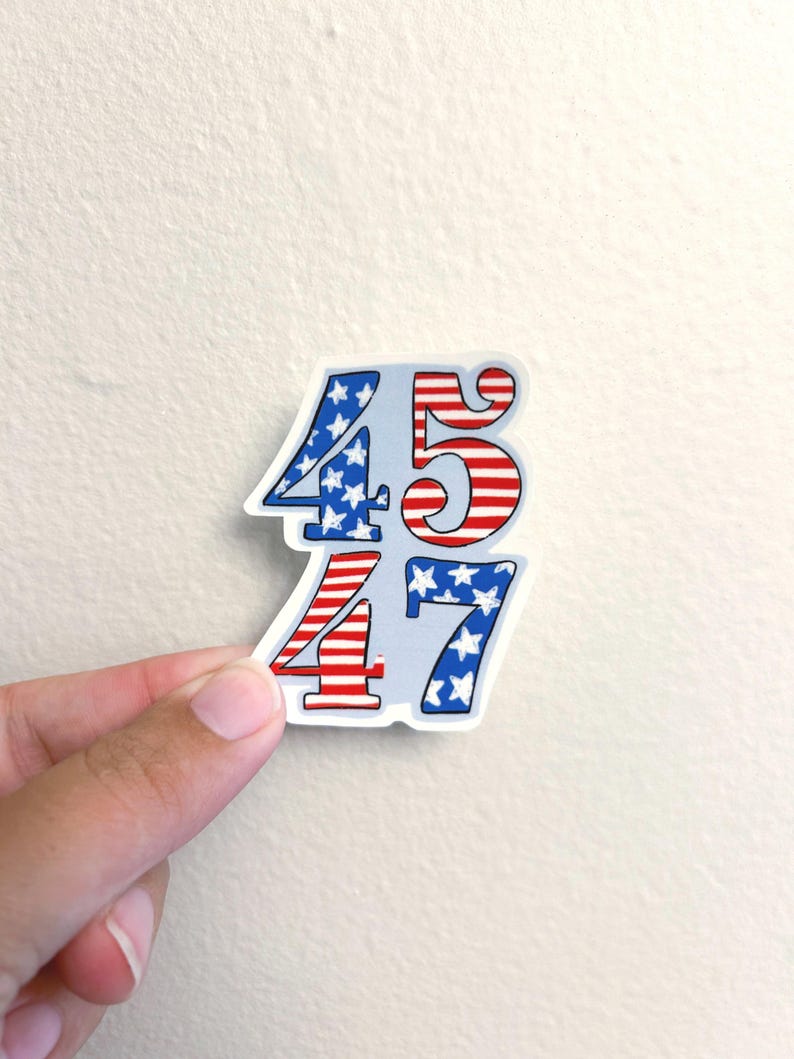 May include: A sticker featuring the numbers 45 and 47, with a patriotic design. The numbers are filled with red and white stripes and blue fields of white stars, reminiscent of the American flag. The sticker is held by a hand.