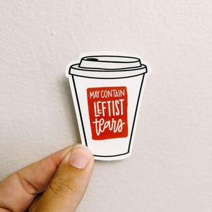 May include: A white sticker in the shape of a coffee cup with a black outline. The sticker has a red rectangle with the words "MAY CONTAIN LEFTIST TEARS" in white. The sticker is held by a hand.
