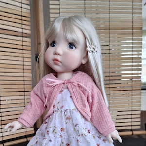 Puede incluir: Una muñeca de porcelana con cabello rubio que lleva un cárdigan rosa y un vestido blanco con flores.