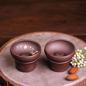 May include: Two brown ceramic tea cups with a floral design inside. Each cup has a perforated sphere at the bottom and sits on a small base. The cups are displayed on a round, light-coloured surface.