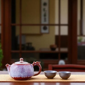 Peut inclure: Un service à thé en céramique. La théière brun-rouge présente un dégradé violet et blanc, un couvercle assorti et une poignée de forme unique. Deux petites tasses à thé brun foncé sont posées à côté de la théière sur un tapis en bambou. L'arrière-plan est flou.