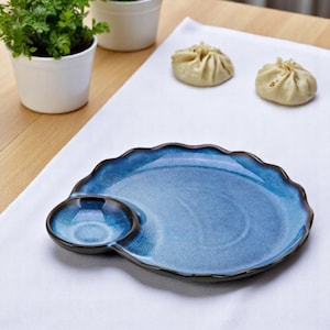 May include: A blue ceramic plate with a wavy edge and an attached small bowl. The plate has a glossy finish and a dark brown rim. Two small, white potted plants and two dumplings are in the background.