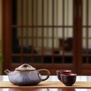 Może przedstawiać: Ceramiczny zestaw do herbaty składający się z czajniczka i dwóch filiżanek. Czajniczek jest w kolorze głębokiego niebieskiego z brązowym uchwytem i pokrywką. Filiżanki są ciemnobrązowe z czerwonym wnętrzem, ustawione na bambusowej macie. Tło to rozmyta drewniana konstrukcja.