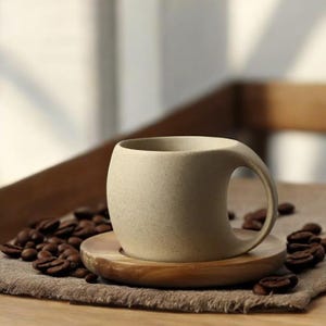 May include: A beige ceramic coffee cup with a unique curved handle sits on a wooden saucer. The saucer is surrounded by coffee beans on a brown fabric surface.