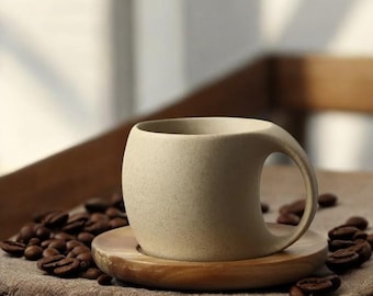 May include: A beige ceramic coffee cup with a unique curved handle sits on a wooden saucer. The saucer is surrounded by coffee beans on a brown fabric surface.